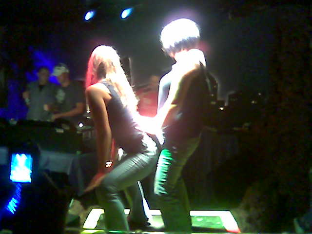 Two people dancing on a stage with colorful lights and a DJ setup in the background