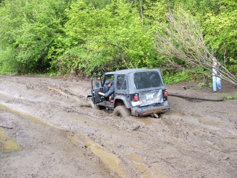 1980 Ford Bronco - A black Jeep Wrangler is stuck deep in mud on a trail, with a person visible inside the vehicle.