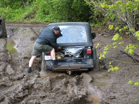 1980 Ford Bronco - A black Jeep Wrangler is stuck in deep mud on a trail, with a person attempting to work on the rear of the vehicle.