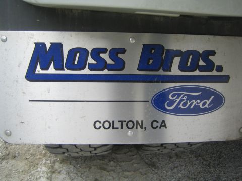 1990 Ford Bronco - brushed-metal dealer license-plate frame reading 'Moss Bros. Ford, Colton, CA' with the blue Ford oval, mounted above dual rear tires.