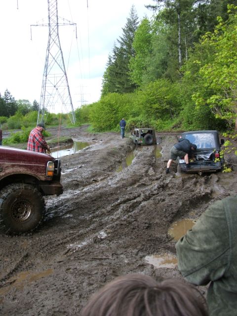 1980 Ford Bronco - Off-road vehicles are stuck in a muddy trail, surrounded by trees and power lines.