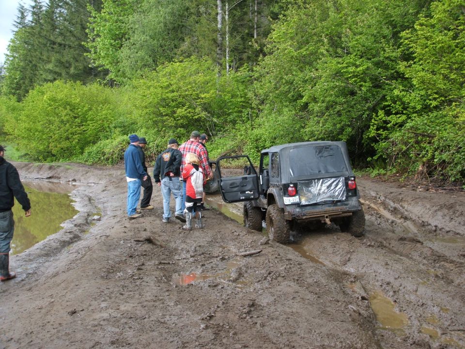 Black soft-top 4x4 stuck in deep mud on a forest trail as a group of onlookers stands nearby