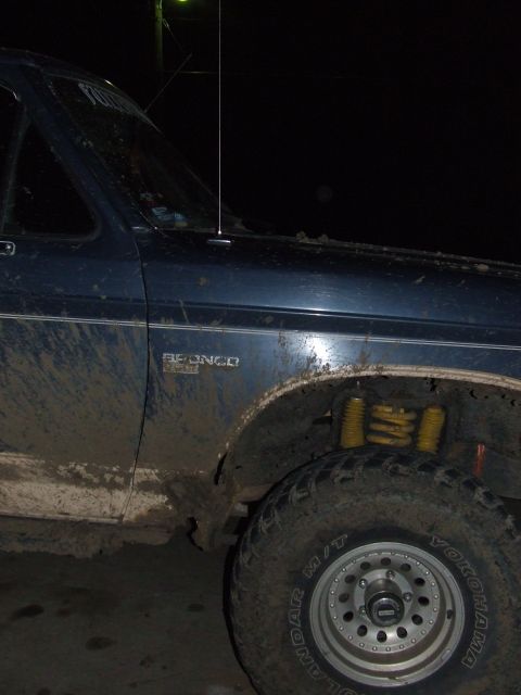 1986 Ford Bronco - Muddy blue Ford Bronco with visible yellow shocks and off-road tires, captured in a low-light, side-angle view.