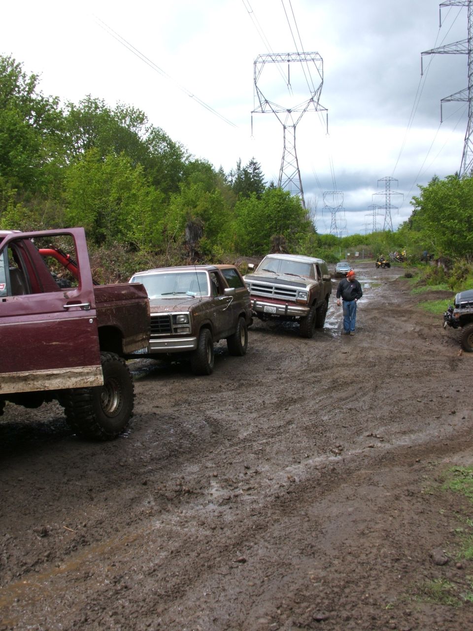 Muddy trail under power lines with 1980s Ford Bronco, Dodge Ramcharger, and lifted flatbed truck