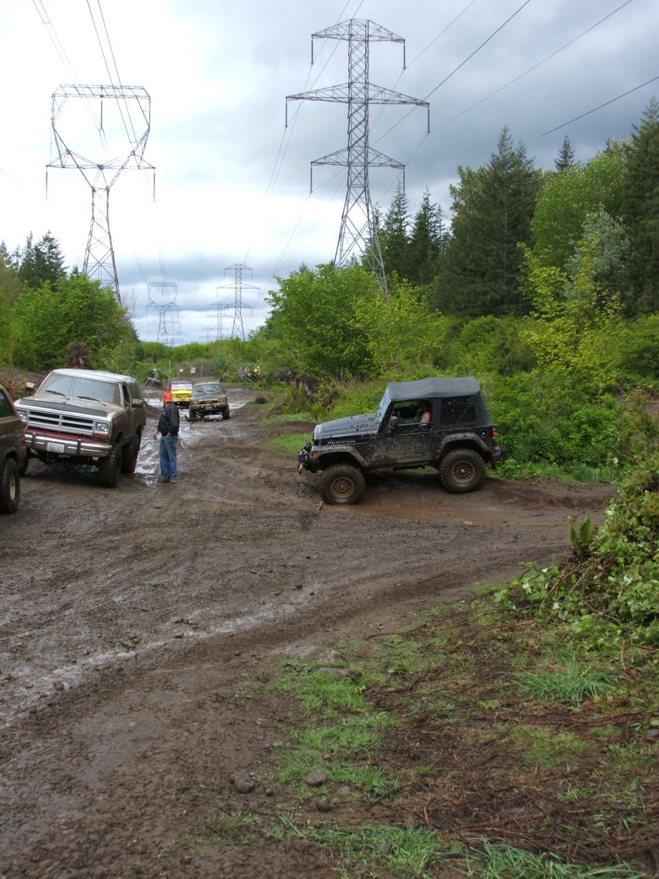 Muddy tan Ford Bronco parked on a wet off-road trail with a black Jeep and power towers behind.