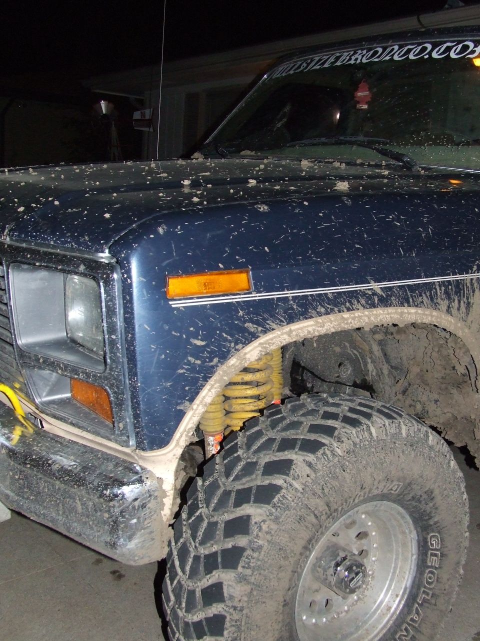 Mud-covered 1986 Ford Bronco front fender, headlight, and tire. Yellow shock absorber visible