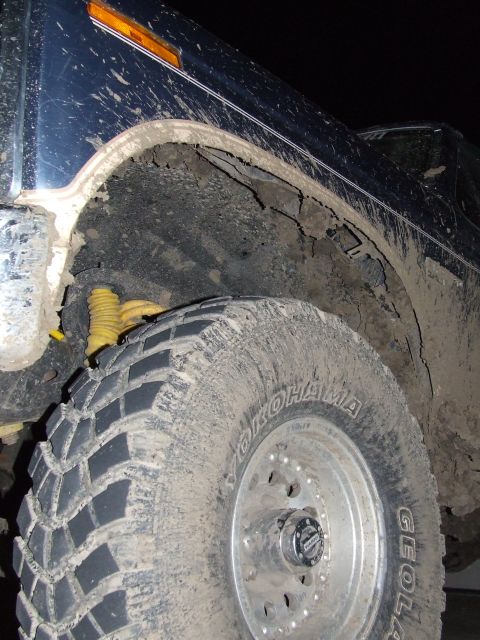 1986 Ford Bronco - Close-up of a mud-splattered 1986 Ford Bronco's wheel well and tire, revealing a yellow shock absorber.