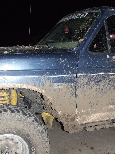 1986 Ford Bronco - A muddy blue 1986 Ford Bronco is shown from a 3/4 front angle, highlighting its lifted suspension and off-road condition.