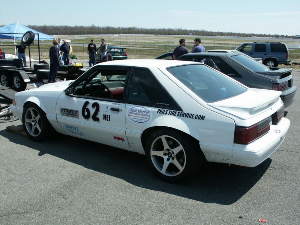 White Ford Mustang with number 62 on the side, parked on asphalt with a trailer and other cars nearby