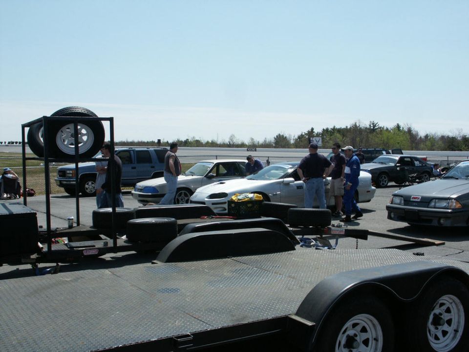 Race track scene with several cars, including a silver Camaro and a grey Ford Mustang, and people gathered around