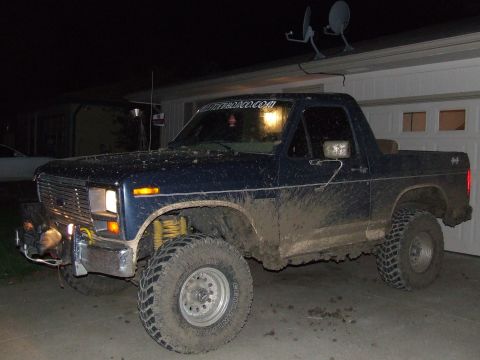 1986 Ford Bronco - This heavily muddied blue Ford Bronco is parked in front of a garage at night.