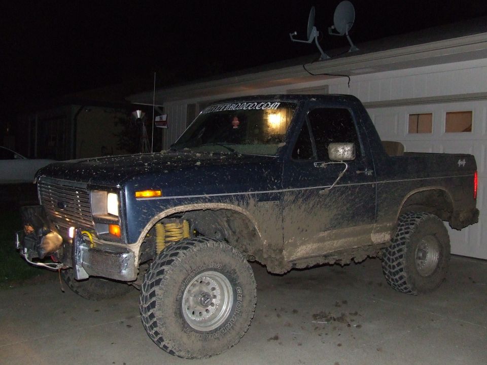 Mud-covered blue Ford Bronco with aftermarket bumper and winch parked in front of a garage