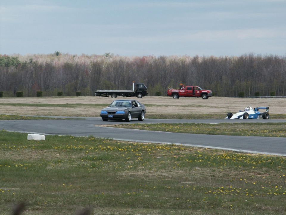 Race track scene with a dark coupe, a flatbed truck, a red pickup truck, and a formula race car