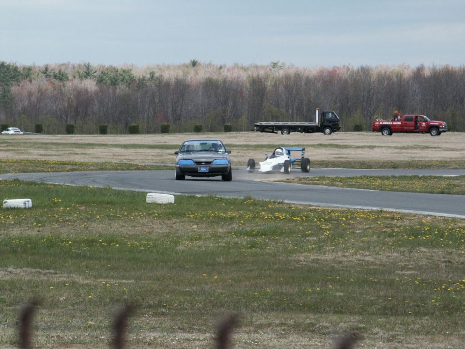 Blue Ford Mustang on a race track with a white open-wheel car. Flatbed truck and red pickup truck in background