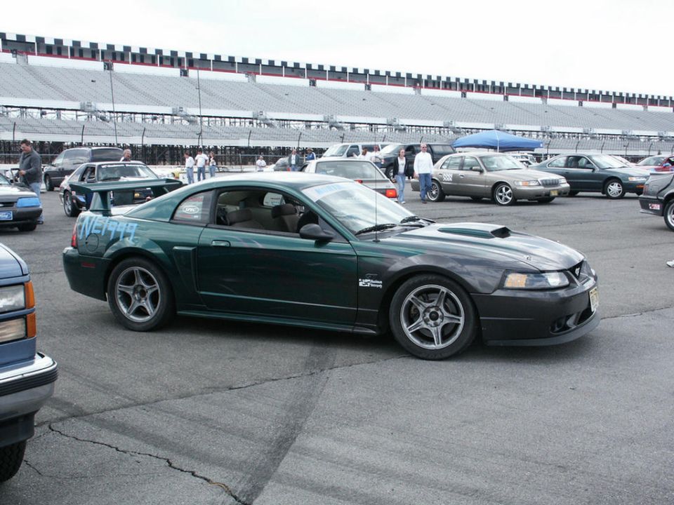 Green Ford Mustang GT with a hood scoop and carbon fiber hood, parked at a racetrack. Text 'NE1999' is spray-painted on the rear quarter panel