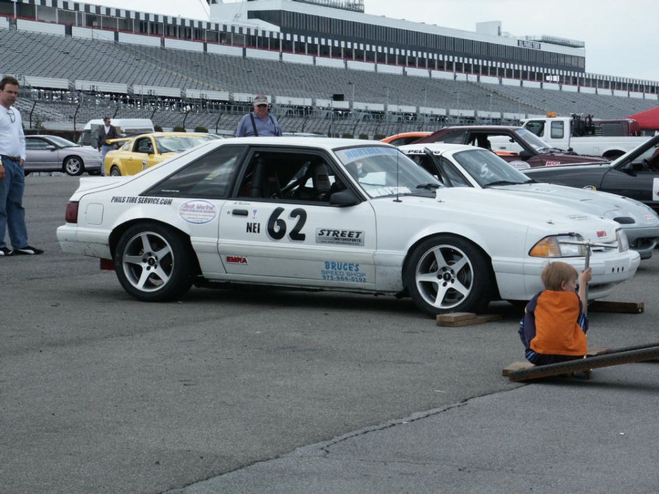 White Ford Mustang with number 62 on the side, parked at a racetrack with grandstands in the background