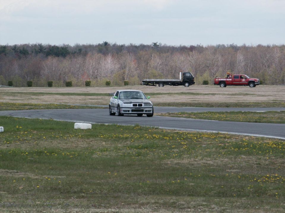 BMW E36 coupe on a race track with a flatbed truck and a red pickup truck in the background