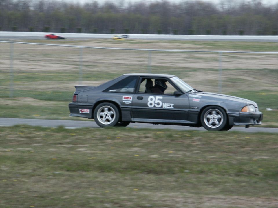 Gray Ford Mustang with number 85 on the door, on a race track. Another red car is visible in the background