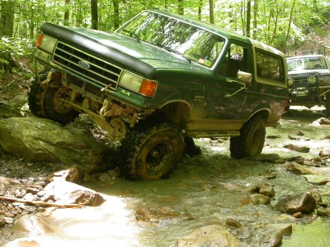 1990 Ford Bronco - A 1990 Ford Bronco tackles a challenging off-road course, splashing through a rocky stream with another vehicle following.