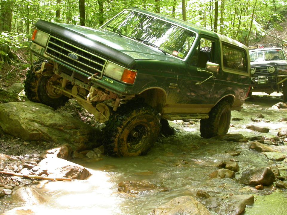 Green 1990 Ford Bronco navigating rocky stream, another SUV behind
