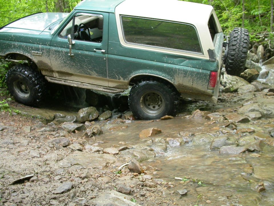 Green Ford Bronco with white top driving through a rocky stream