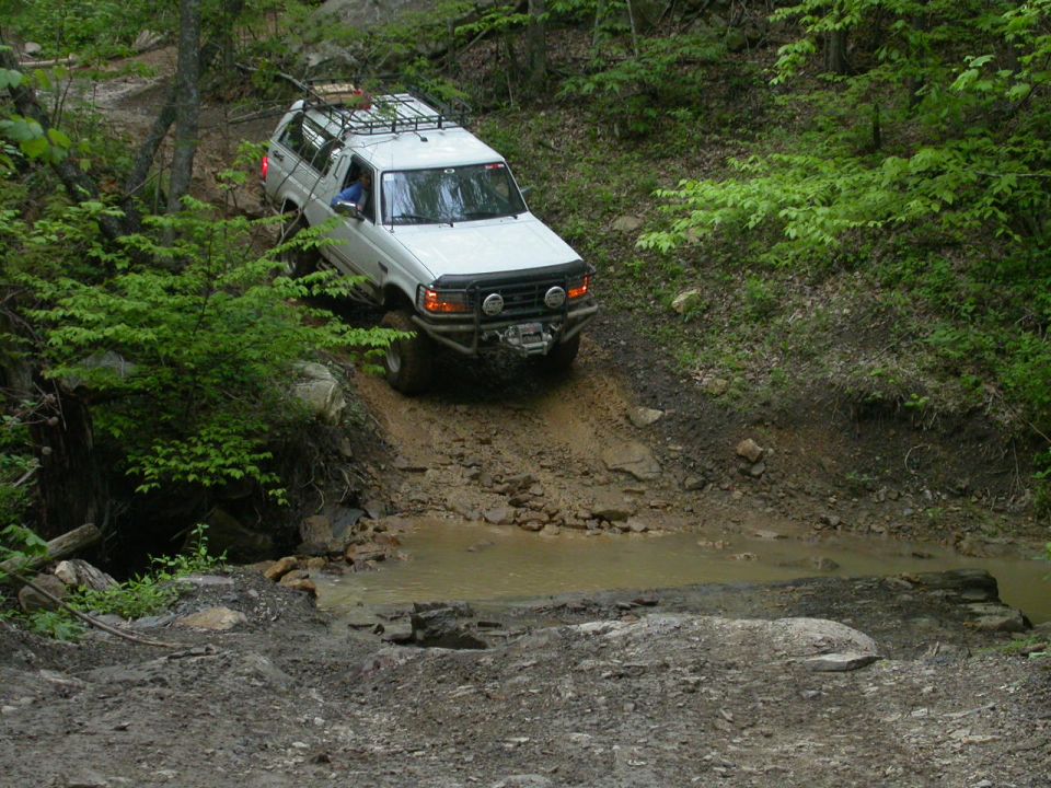 1990 Ford Bronco navigating a muddy off-road trail with trees on either side