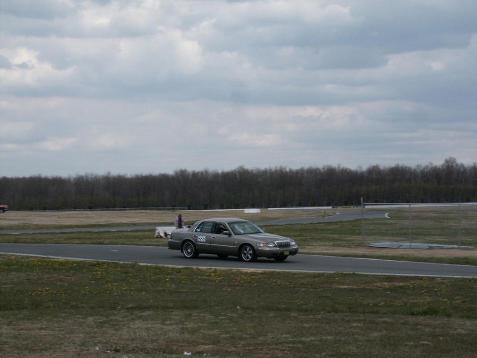 Gray 2001 Mercury Grand Marquis driving on a paved racetrack with a cloudy sky