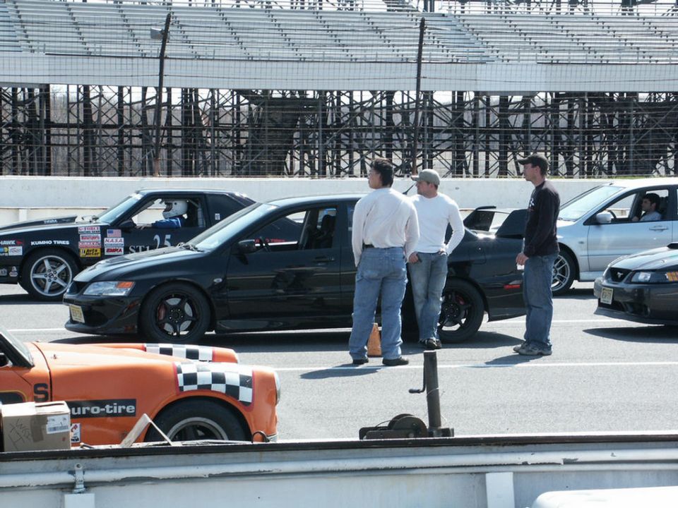 Race track scene with several cars and people standing around. An orange car is in the foreground