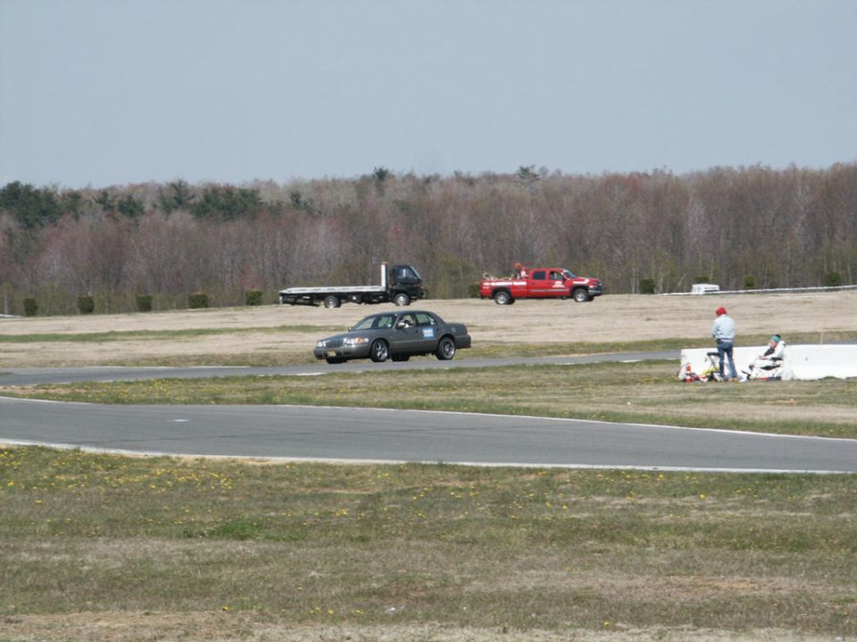 Dark gray 2001 Mercury Grand Marquis on a race track, 3/4 front view, with a flatbed truck and a red pickup truck in the background