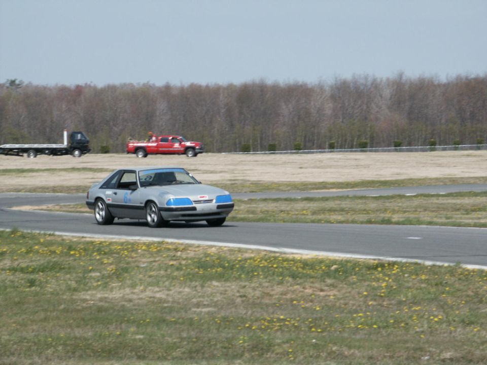 Silver coupe with blue accents on front bumper driving on a race track. Red pickup truck and flatbed trailer in background