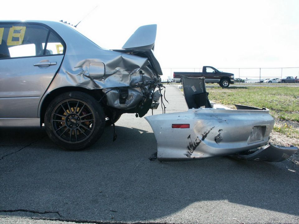 Silver Mitsubishi Evo VII with rear-end damage, bumper detached. Other cars and a truck in background