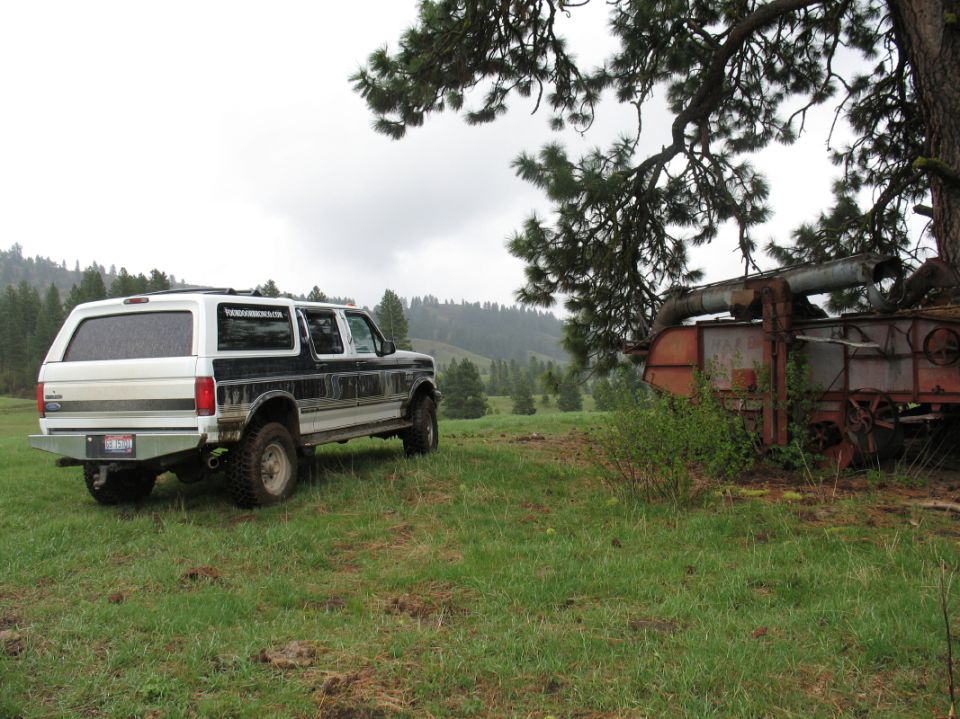 Muddy white four-door Ford Bronco conversion parked in a meadow near rusty farm machinery