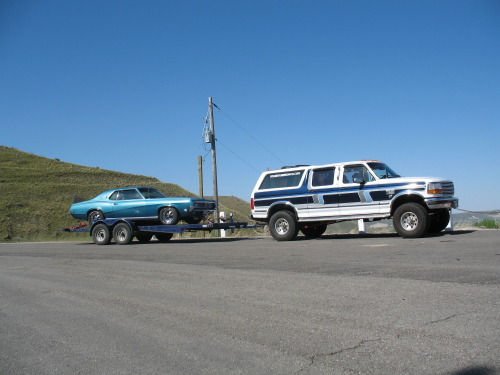 White/blue lifted SUV towing trailer with blue vintage muscle car on a paved lot.