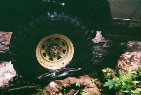 1978 Ford Bronco - A close-up view of a large off-road tire and wheel, partially submerged in water and surrounded by rocks.