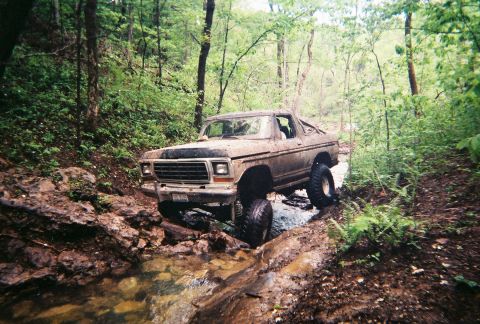1978 Ford Bronco - A lifted Ford F-Series truck navigates a rocky, wooded stream bed.