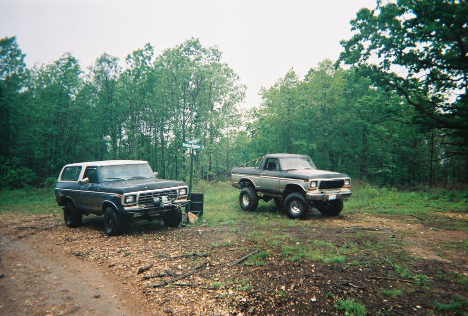 Dark blue and white 1978 Ford Bronco parked on a dirt road next to a lifted pickup truck, with trees in the background