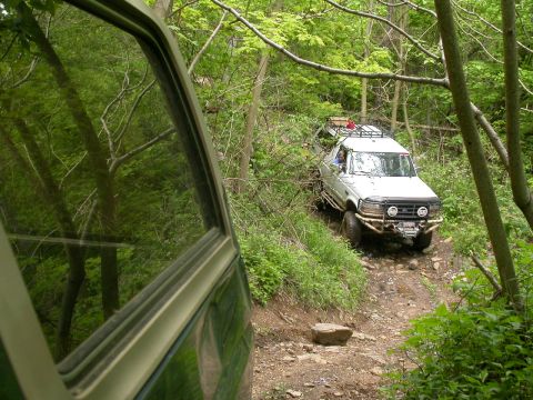 1990 Ford Bronco - A white 1990 Ford Bronco navigates a challenging off-road trail through a dense forest.