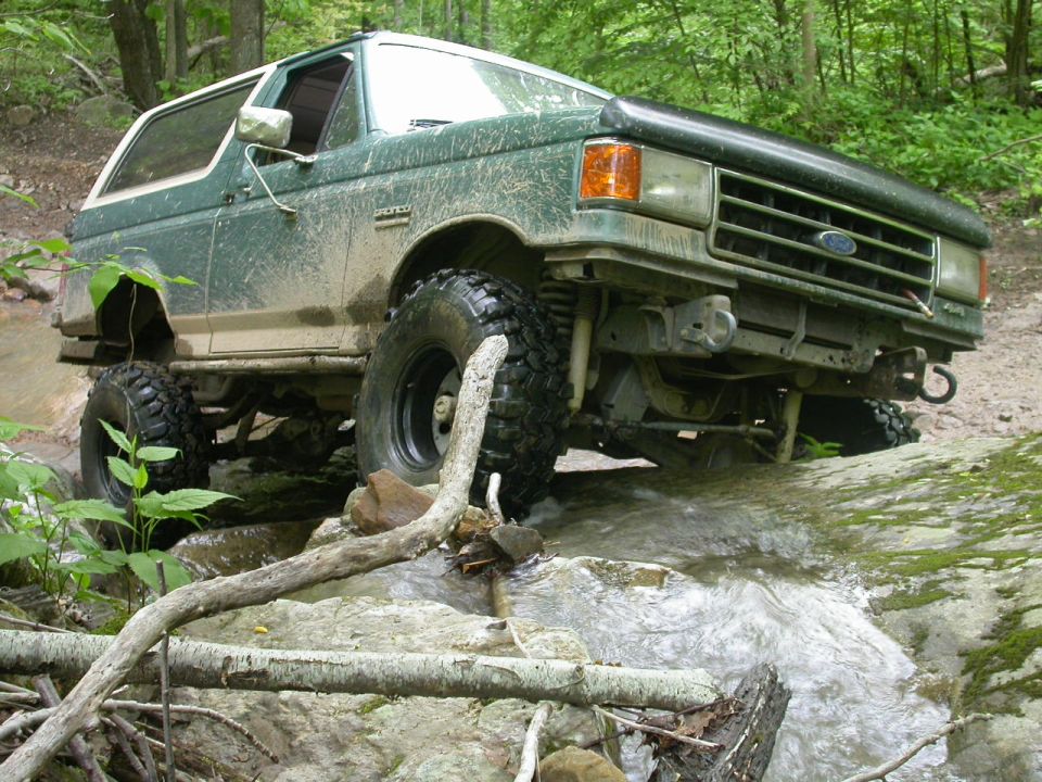 1990 Ford Bronco driving through a rocky stream, covered in mud