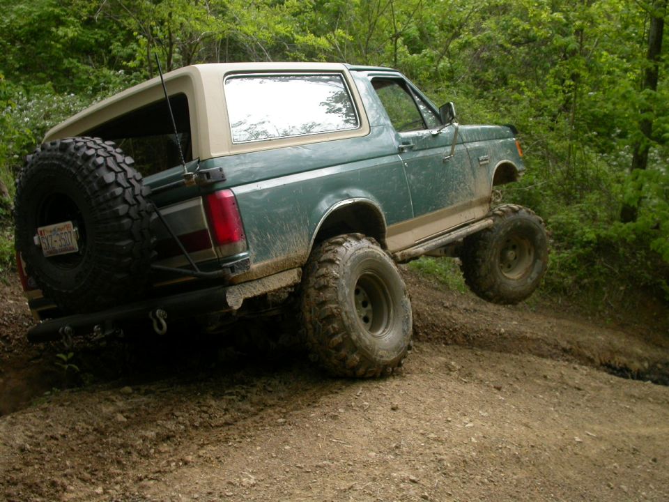 Green Ford Bronco with tan top, lifted suspension, and spare tire on rear, driving on dirt trail