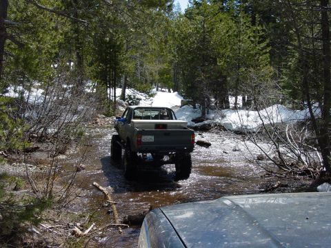 1986 Ford Bronco - Two vehicles navigating a stream in a wooded area with snow patches.