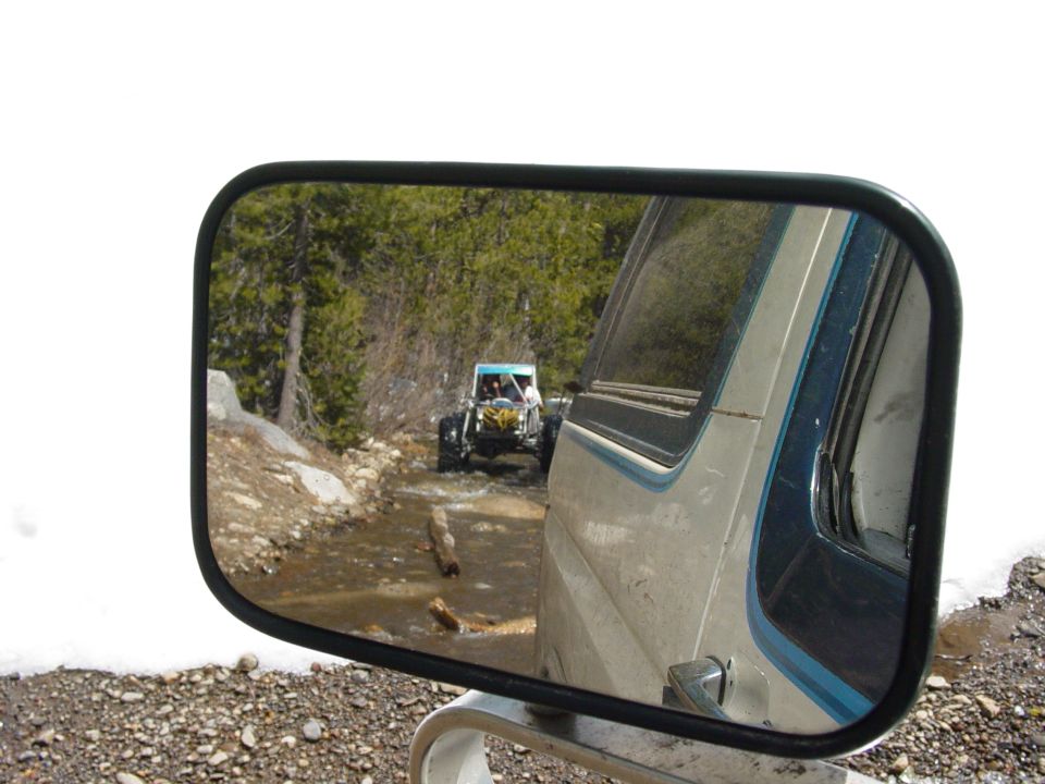 Side mirror view of a buggy driving on a dirt road with trees and snow