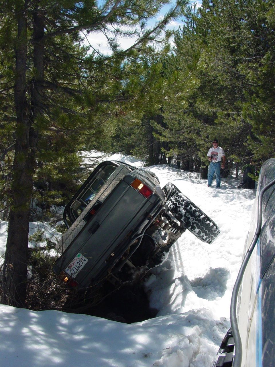 Gray SUV tilted in snow with one wheel in the air, a person stands in the background