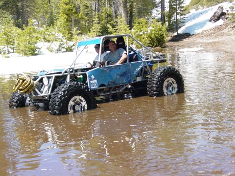 1986 Ford Bronco - A custom buggy navigates through shallow water, with trees and snow-capped hills in the background.