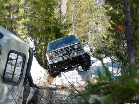 1986 Ford Bronco - A blue Ford Bronco navigates a rocky, off-road trail with a second vehicle partially visible on the left.