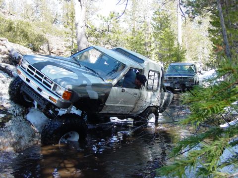1986 Ford Bronco - A camouflaged 1986 Ford Bronco navigates a water obstacle, followed by another truck in the woods.