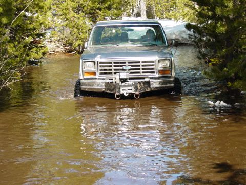 1986 Ford Bronco - Ford Bronco navigates a water crossing in a wooded area with snow patches visible.