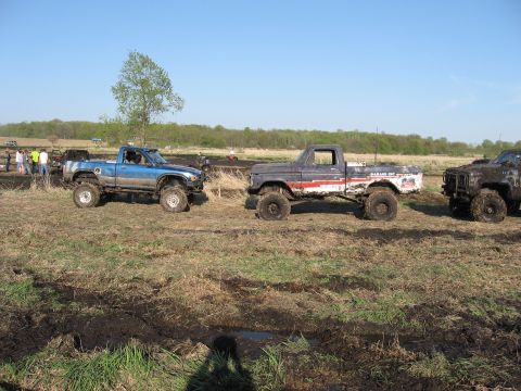 1977 Ford F-150 - Several modified trucks are gathered in a muddy outdoor area, likely for an off-road event.