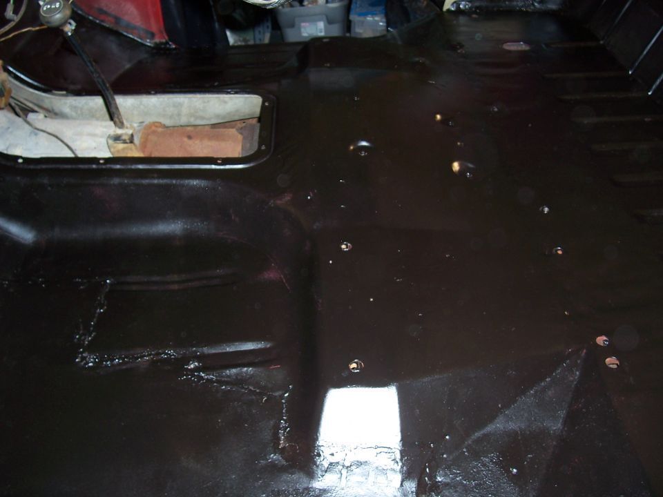 Vehicle floor pan with black coating, showing transmission tunnel opening and some rust spots