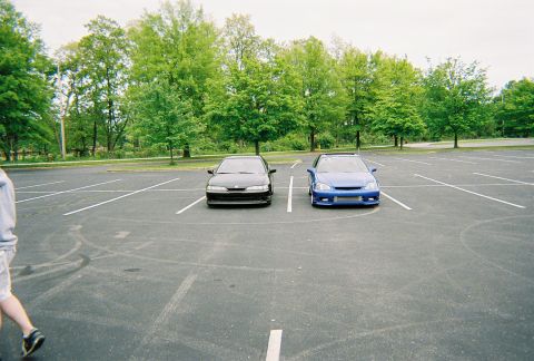 1991 Ford Probe - Two tuner cars are parked side-by-side in an empty parking lot with trees in the background.