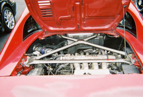 1991 Ford Probe - Engine bay of a red Toyota MR2, showing the engine and cross brace. A portion of another vehicle is visible to the left.